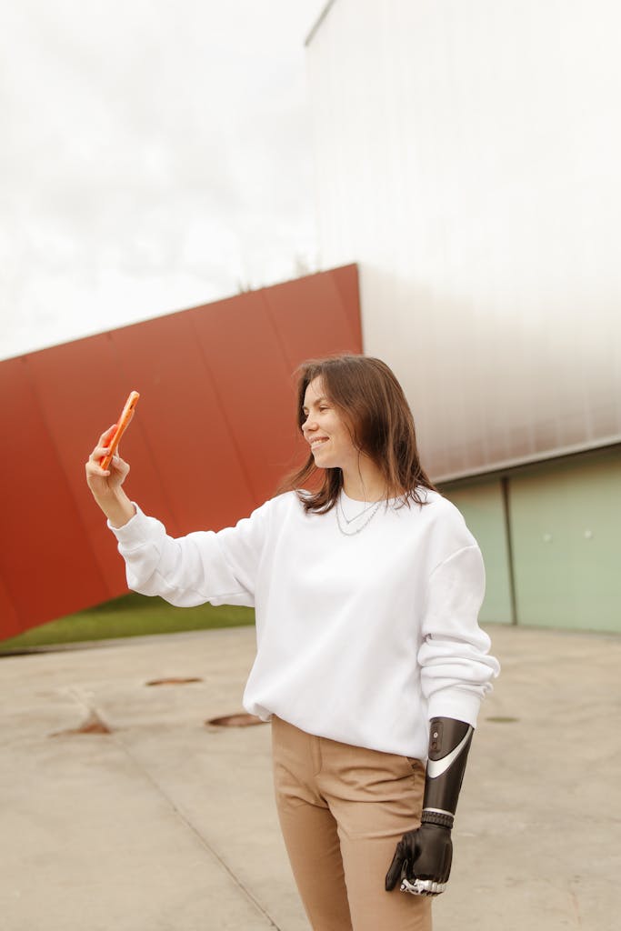 Smiling woman with prosthetic arm taking a selfie outside against a modern building backdrop.