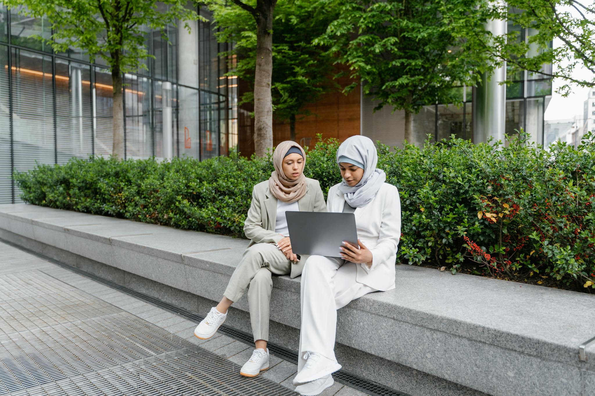 Two Muslim women in hijabs working together on a laptop outdoors in a modern park setting.