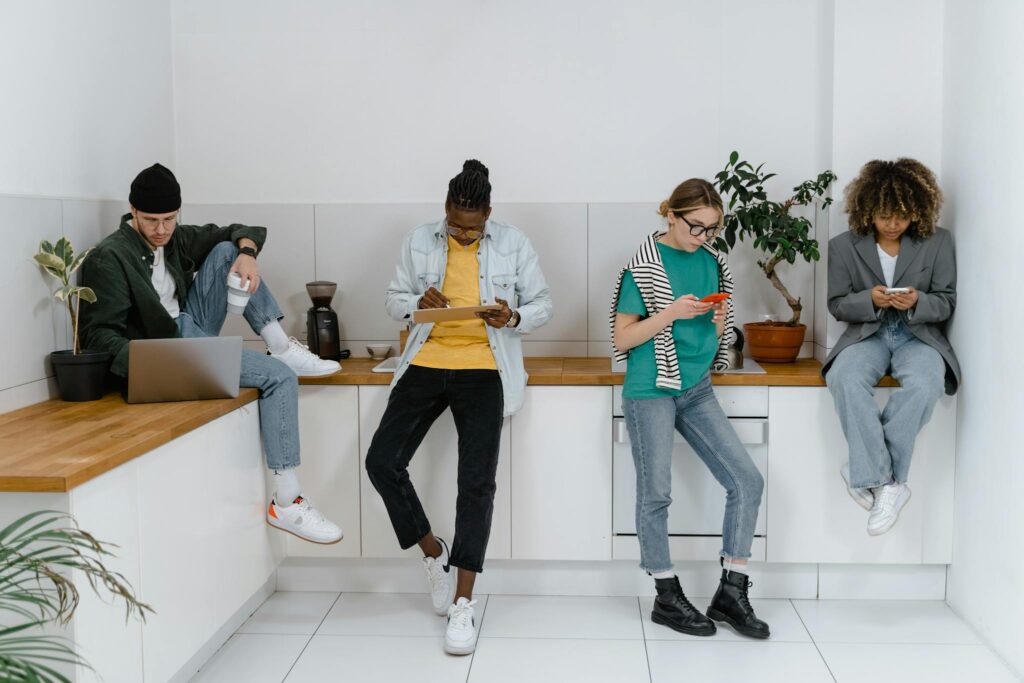 Young adults on digital devices in a modern office kitchen area, emphasizing diversity and teamwork.
